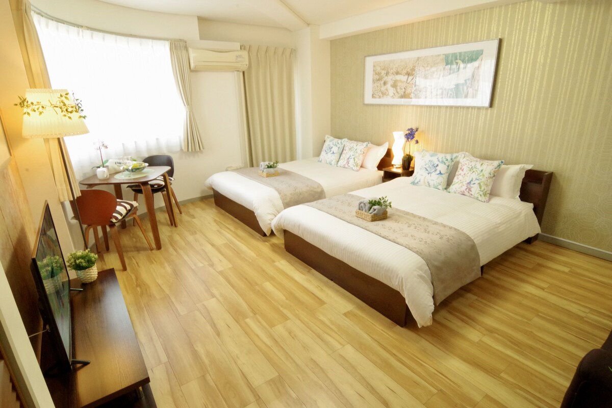 A hotel room with two beds, a desk with a chair, a small table with a vase, and a window with curtains.