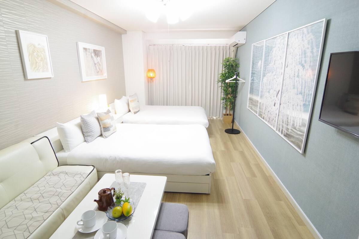 A modern hotel room with a sofa, two beds, a dining table with a tray of lemons, and artwork on the walls.