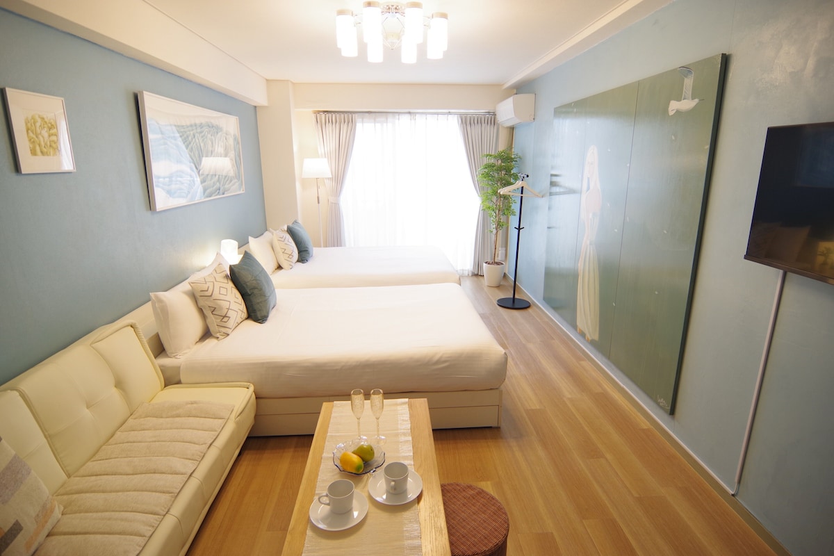 A hotel room with a bed, a sofa, a small table with a tray of fruit and glasses, and a TV on the wall.