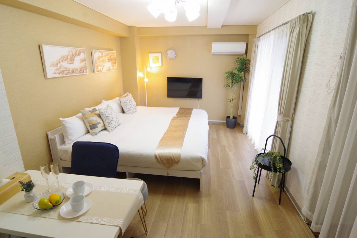 A hotel room with a bed, bedside table with a fruit bowl, a TV, a potted plant, and a window with curtains.