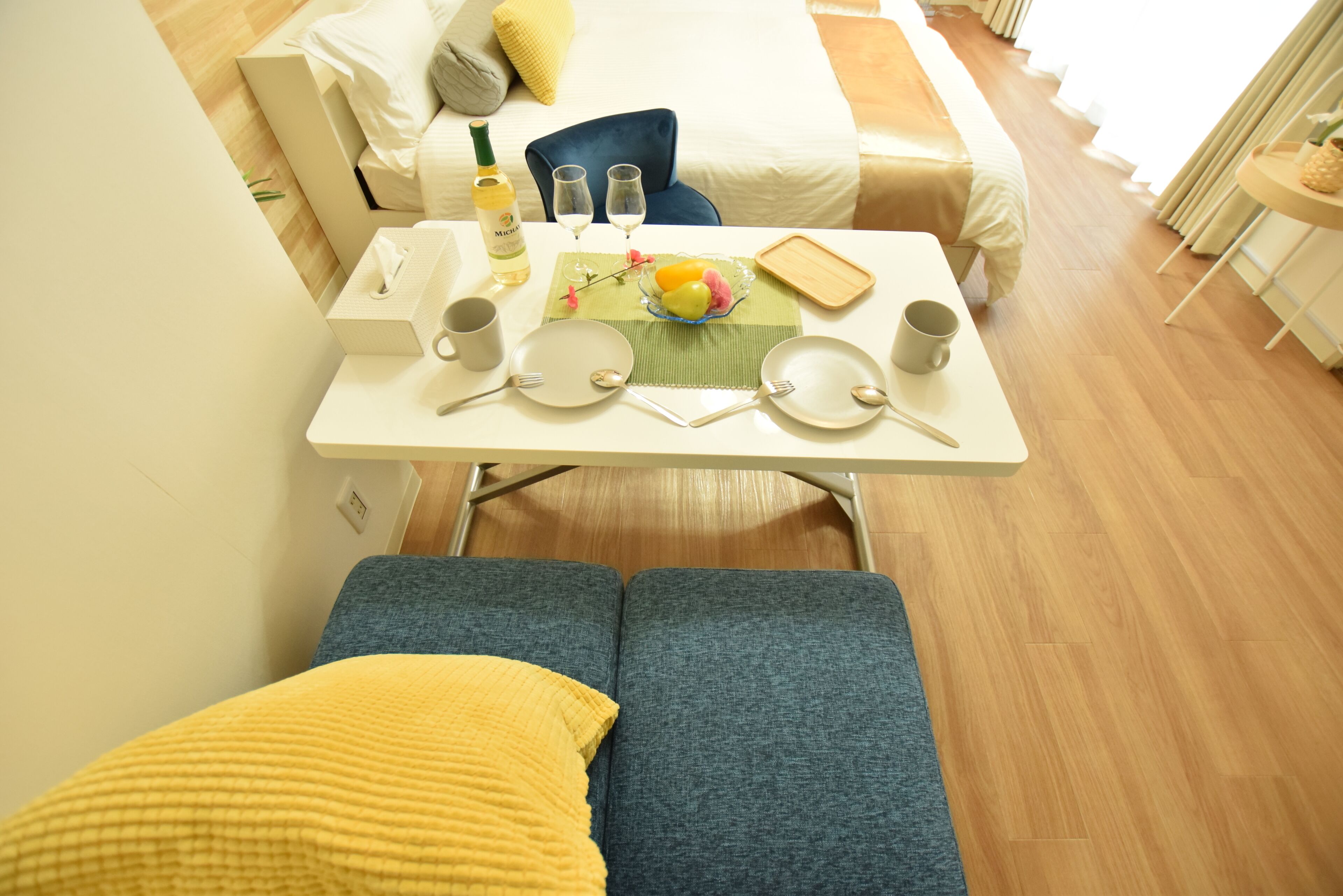 A neatly arranged breakfast table with plates, cutlery, and a bottle of wine, set in a modern hotel room with a bed and a blue chair in the background.