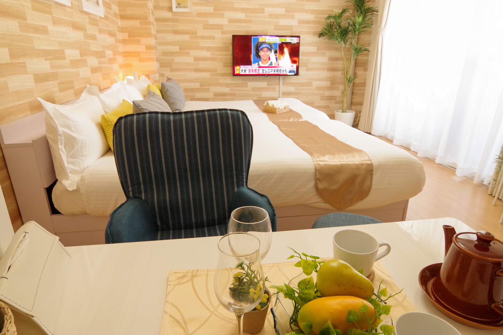 A hotel room with a bed, a striped armchair, a table with a teapot, a cup, and a bowl of fruit.