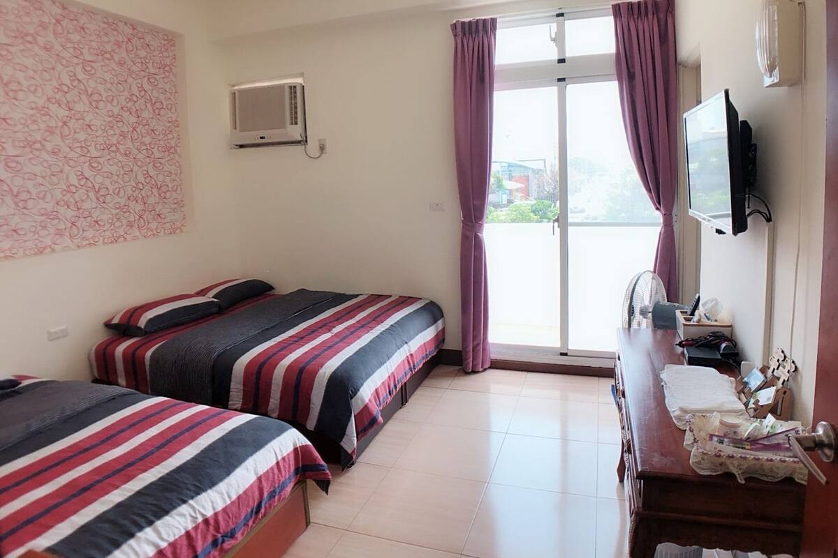 Habitación de hotel con dos camas, televisor, escritorio con ventilador y ventana con cortinas.