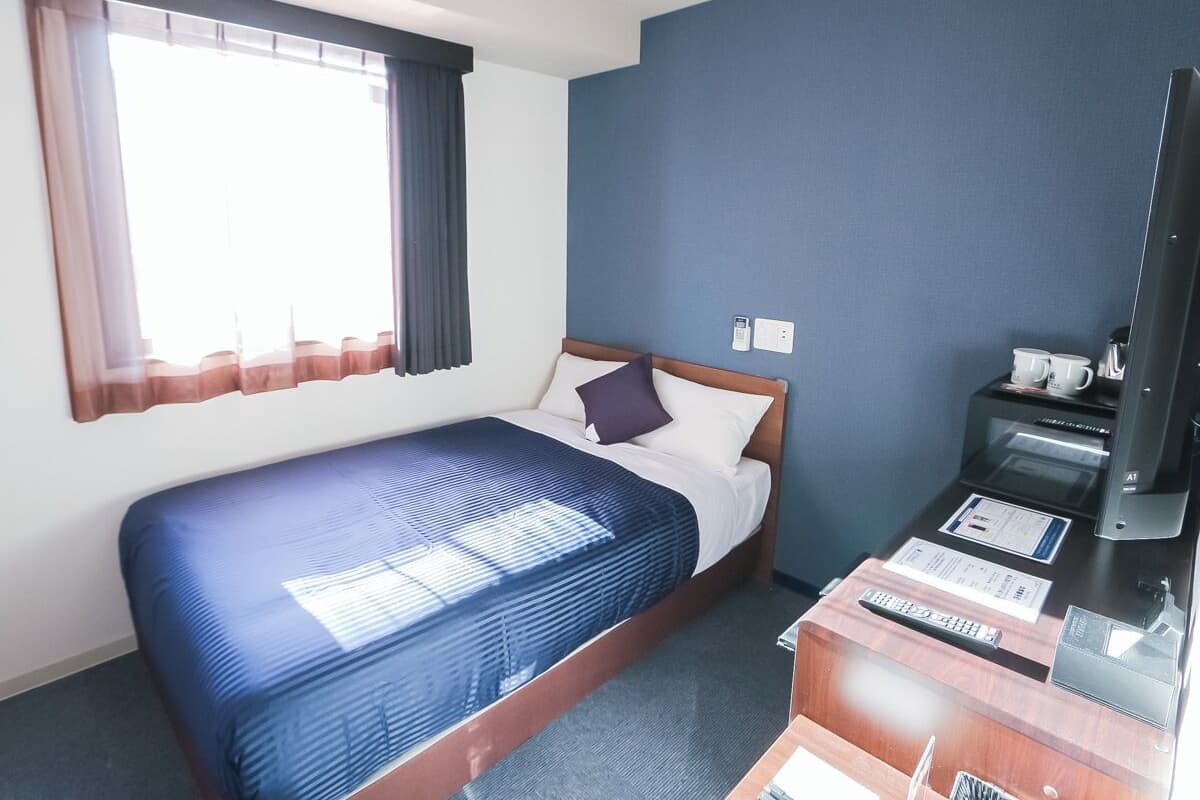 A hotel room with a bed, a desk with a computer, a telephone, and a window with curtains.