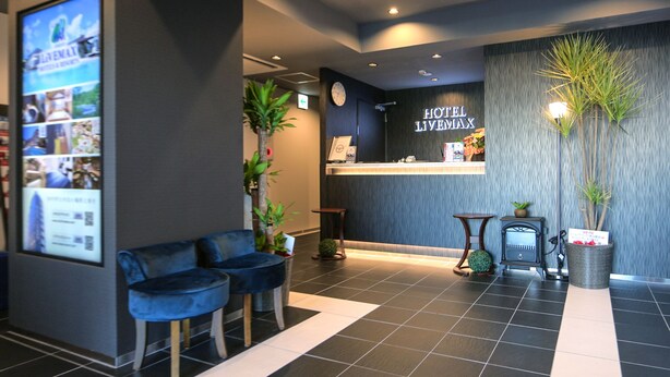 Ein Hotel-Empfangsbereich mit einer Rezeption, zwei blauen Sesseln, einer Uhr und einem an der Wand montierten Bildschirm, der Bilder zeigt.