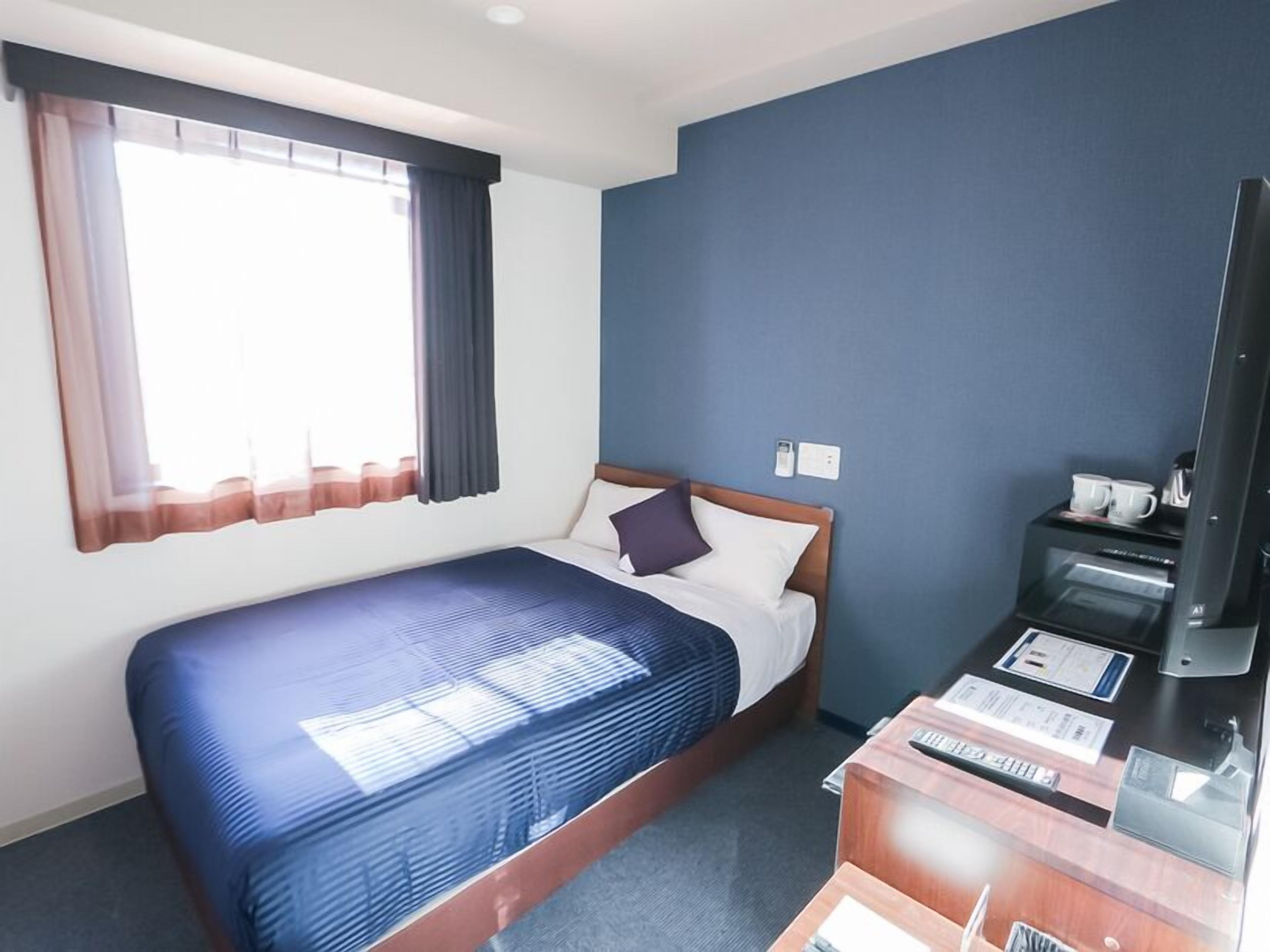 A hotel room with a bed, a television, a desk with a computer, and a window with curtains.