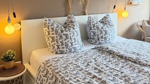 Un lit recouvert d’une couvre-lit à motifs, deux oreillers ornés de motifs de papillons, un luminaire fixé au mur et une petite plante posée sur une table de chevet.