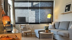 Un salon moderne avec un canapé gris, un fauteuil en osier, une table basse ronde et une grande fenêtre équipée de stores.