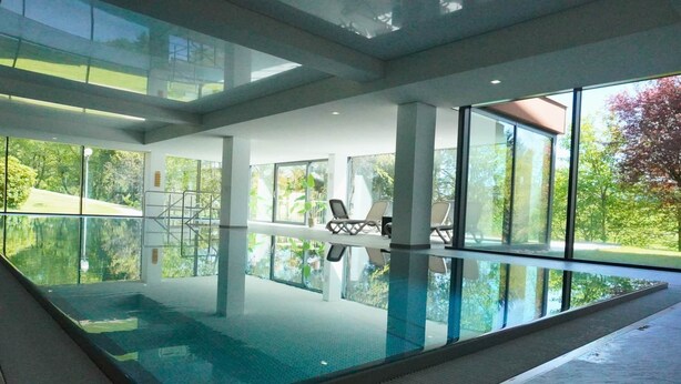 Une piscine intérieure moderne avec des parois en verre transparent, un sol carrelé et une vue sur l’extérieur.