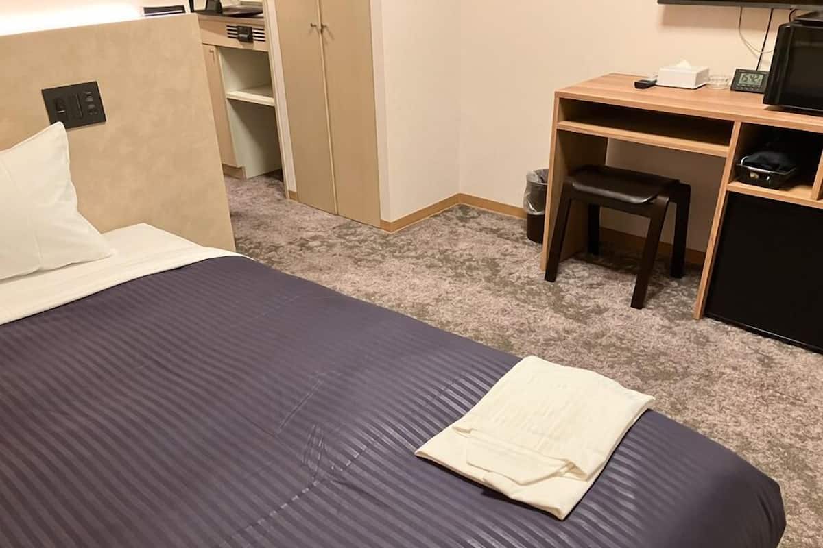 Un lit bien fait, recouvert d’une couvre-lit bleu foncé, avec un oreiller blanc et un drap blanc plié. On y trouve un bureau en bois avec une chaise, une petite poubelle et un téléviseur posé sur un meuble.