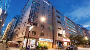 Un hotel de varios pisos con un cartel llamativo que dice 'HOTEL LIVEMAX', una entrada visible, máquinas expendedoras y un área de estacionamiento con un automóvil.