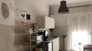 A kitchen with white cabinets, a stainless steel refrigerator, and a white countertop. There is a hanging light fixture and a window with lace curtains. - Casa Isabella (Verona)