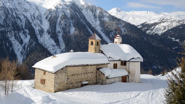 A snow-covered church with a bell tower and a small annex, surrounded by mountains and a clear sky.