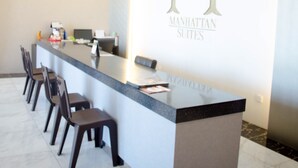 Ein Empfangsbereich mit einer langen Theke, mehreren Sesseln und einer Wand mit dem großen 'M'-Logo und dem Schriftzug 'MANHATTAN SUITES'.