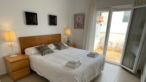 A neatly made bed with white linens and decorative pillows, a wooden nightstand with a lamp, and a view of a balcony through a sliding glass door.