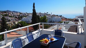 A balcony with a table set for two, overlooking a coastal town with white buildings and a clear blue sky.