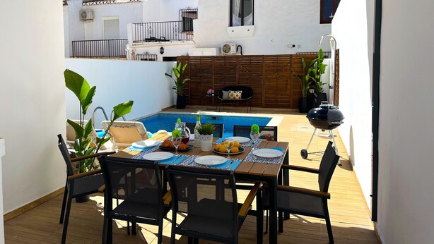 A covered outdoor dining area with a pool, wooden deck, and a view of a building with a balcony.