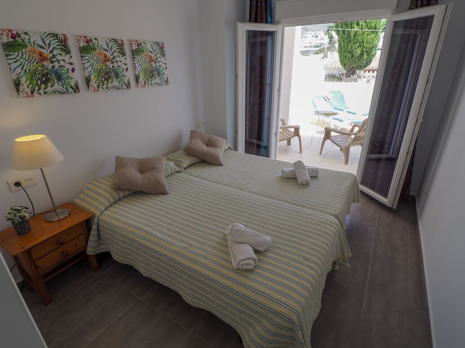 A bedroom with two beds, a nightstand, a lamp, and a view of an outdoor patio with chairs and a table.