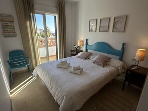 A neatly made bed with pillows and towels, a nightstand with a lamp, a chair, and a balcony with curtains.