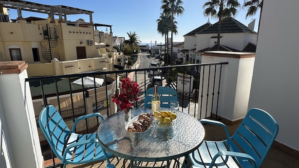 A balcony with a round glass table, blue chairs, a vase with red flowers, and a bottle of wine. - Nerja Paradise Rentals - Apartment Chimenea A1E (Nerja)