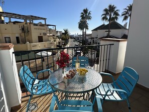 Un balcon avec une table ronde en verre, des chaises bleues, un vase rempli de fleurs rouges et une bouteille de vin.
