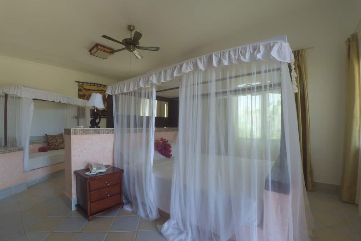 A bedroom with a bed covered by a sheer white curtain, a bedside table with a lamp, and a ceiling fan.