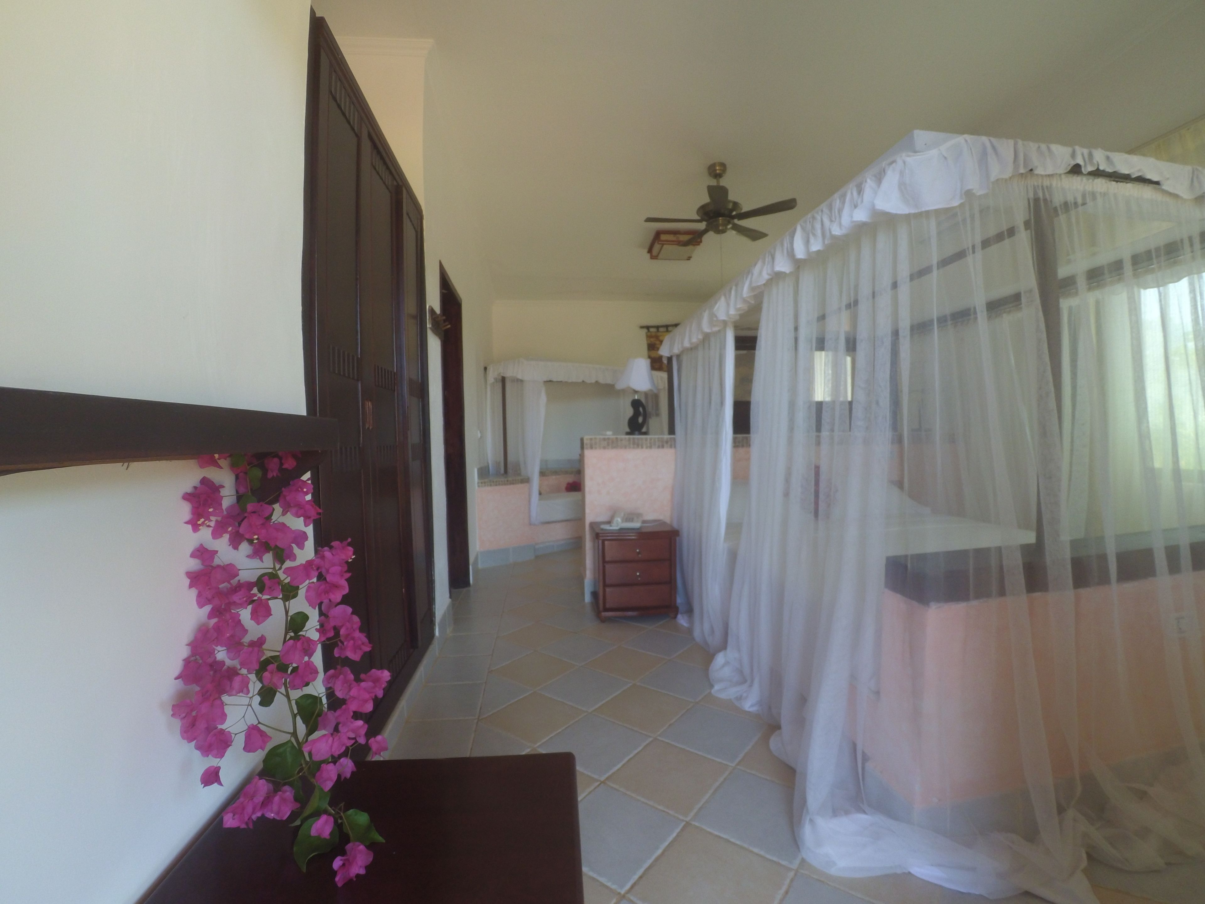 A hotel room with a bed covered in white curtains, a bedside table with a lamp, and a view of the hallway.