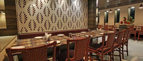 Un restaurante con una larga mesa dispuesta para comer, sillas de mimbre y una pared decorativa con motivos de hojas.