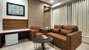 A modern living room with a brown leather sofa, a small coffee table, a white mini-fridge, and a framed abstract painting on the wall. - SVASTI Inn Jamnagar (Jamnagar)