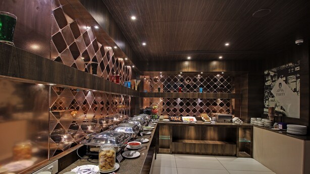 A buffet setup with various food items and utensils, a counter with plates and bowls, and a decorative wall with a geometric pattern. - SVASTI Inn Jamnagar (Jamnagar)