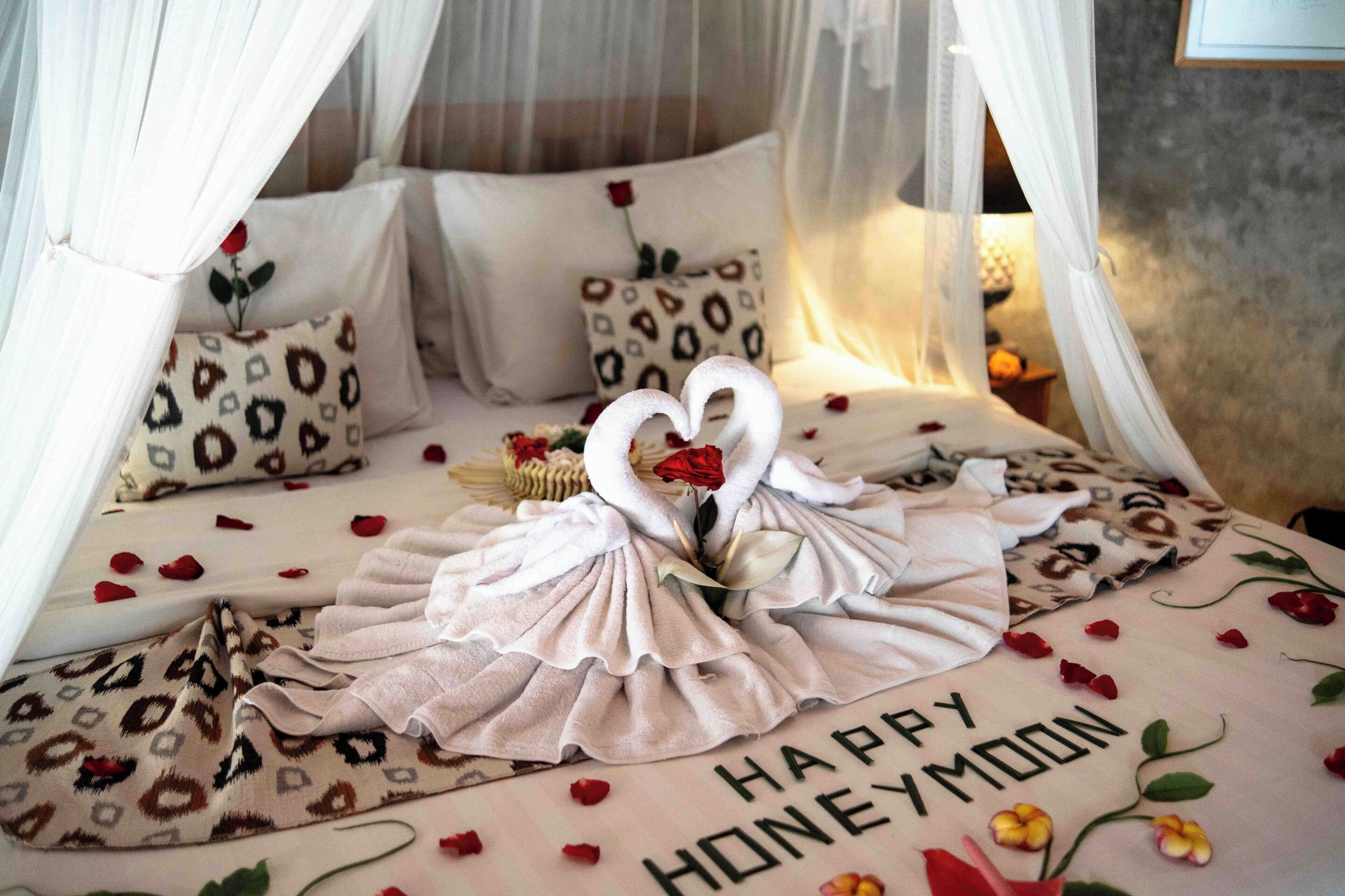 A bed with a canopy, decorated with a heart-shaped arrangement of rose petals and a bouquet of flowers. The bedspread has a message that reads "HAPPY HONEYMOON."