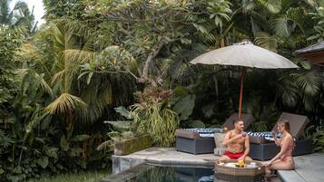A tropical outdoor pool area with lush greenery, a wicker table with a beverage, and lounge chairs.