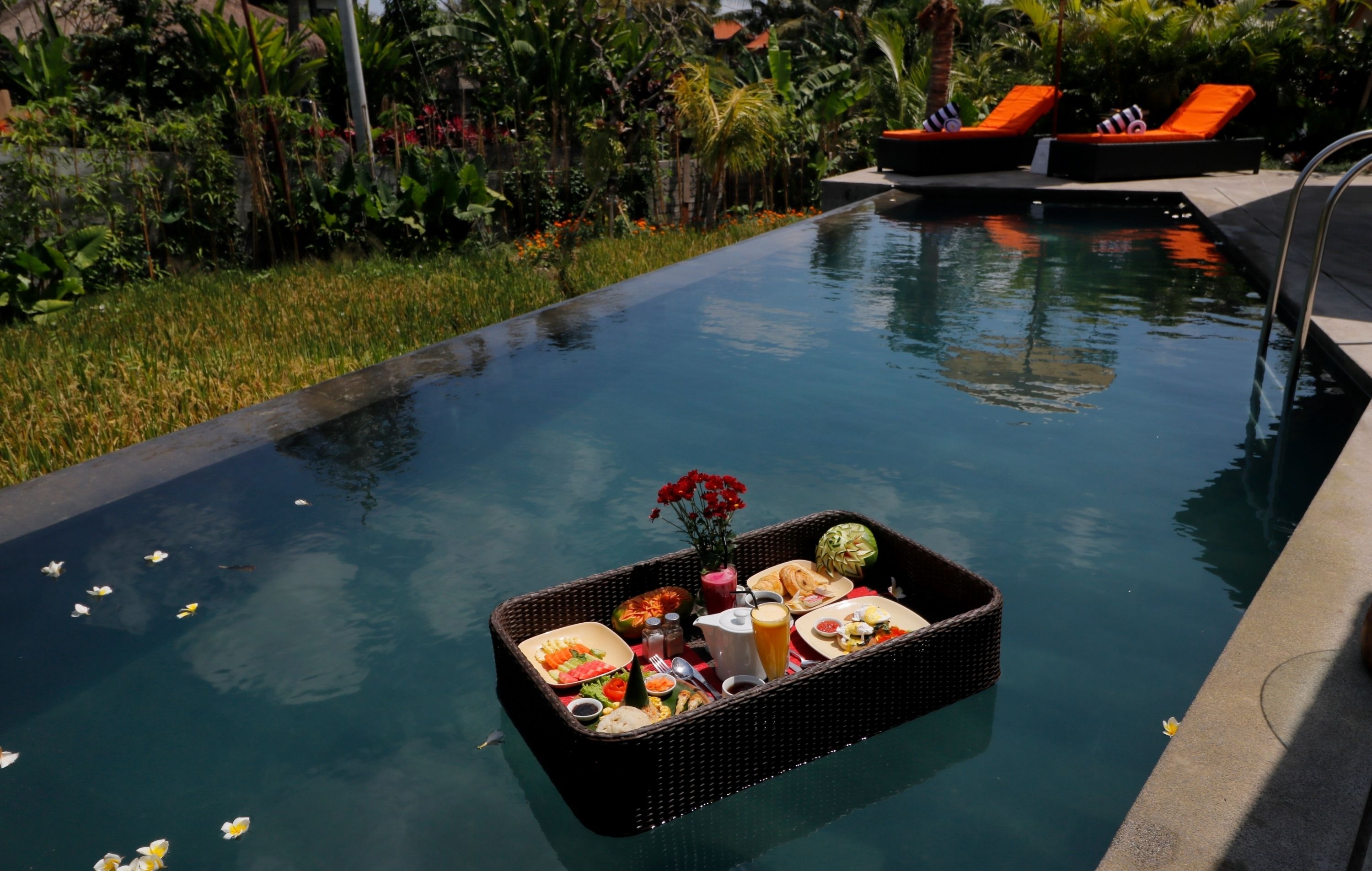 A poolside dining setup with a floating tray of food and drinks, surrounded by lush greenery and lounge chairs.