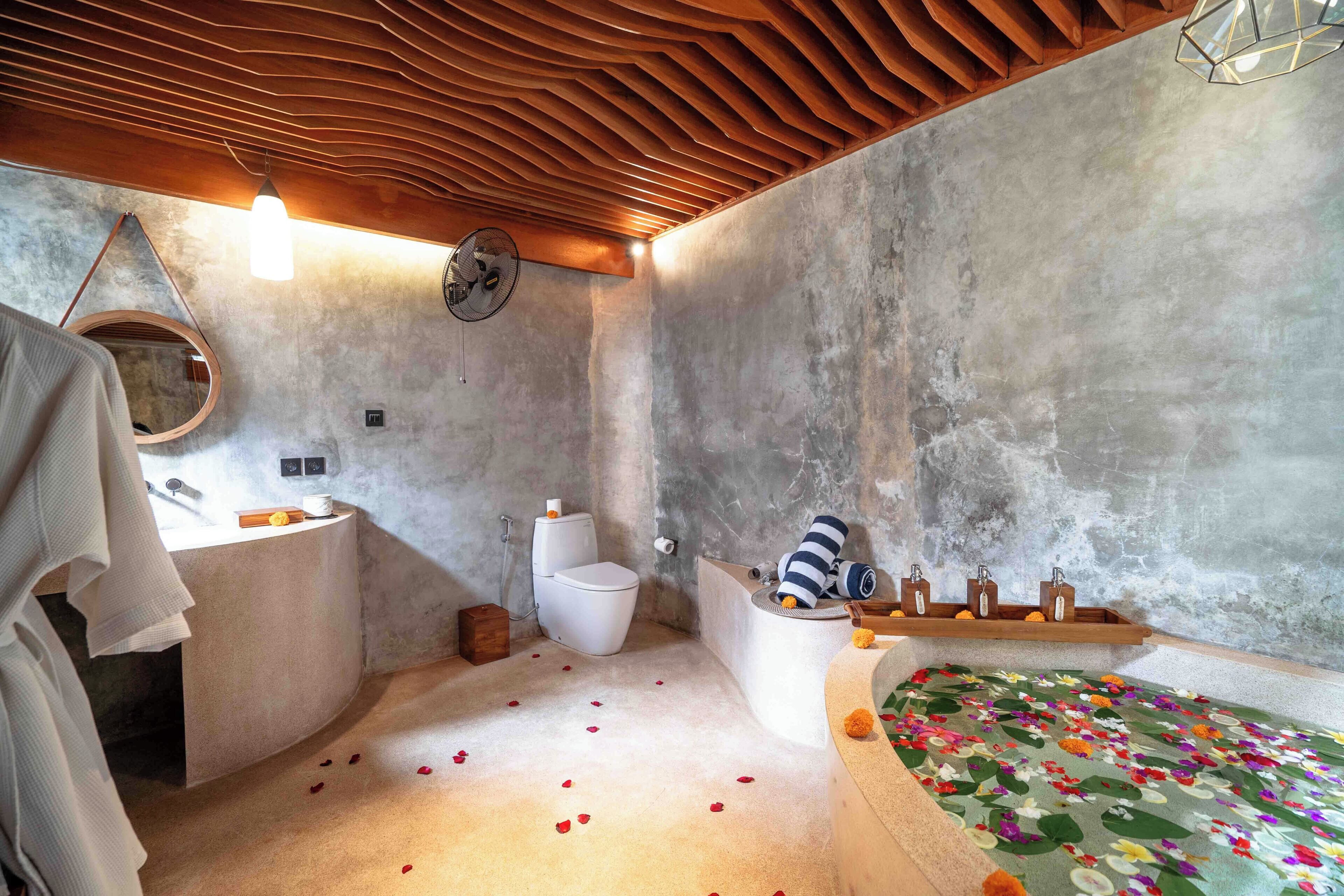 Een badkamer met een houten plafond, een ronde spiegel, een toilet, een wastafel en een bad met een badmat met bloemenmotief.