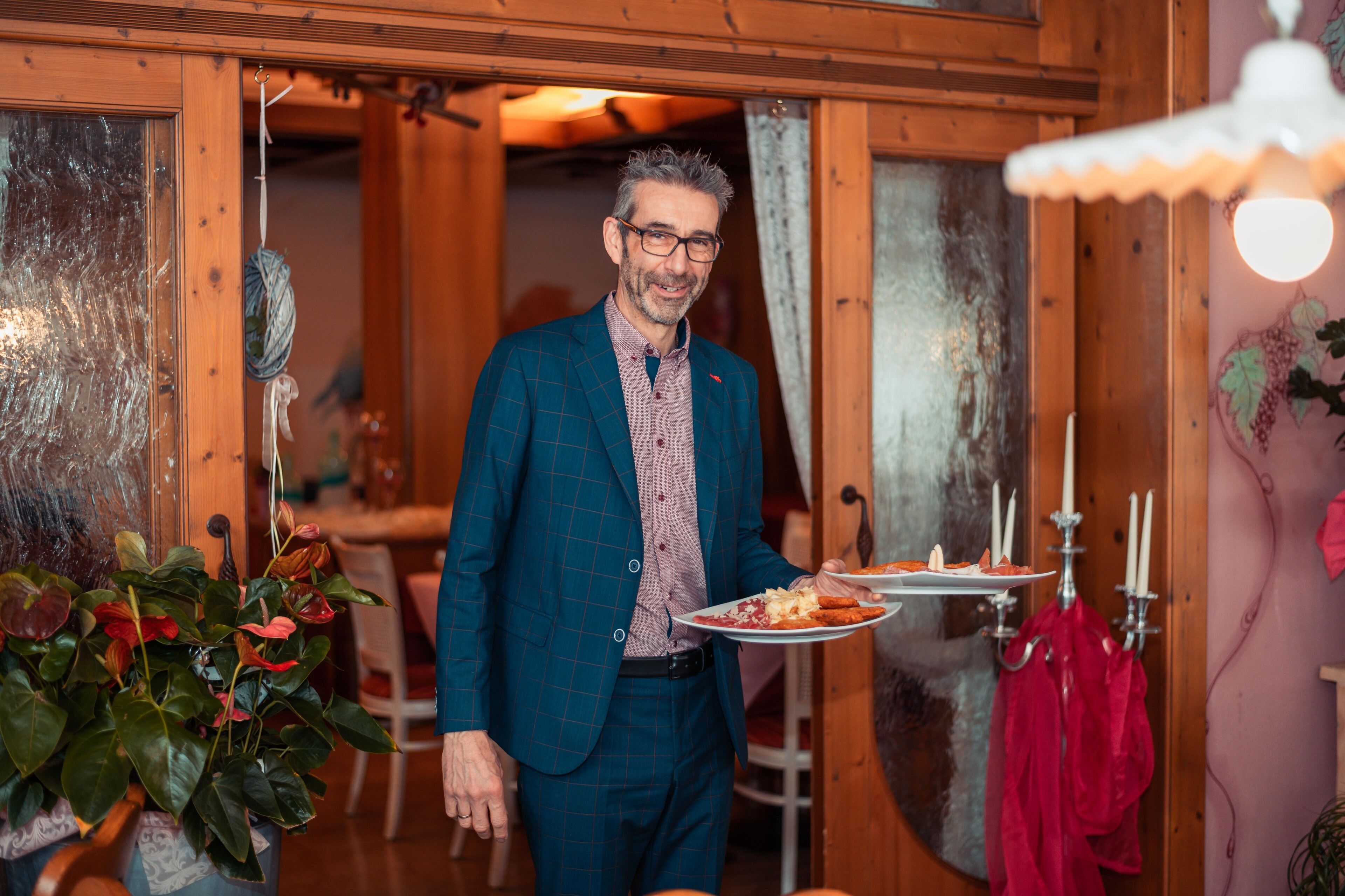 Ein Mann in einem blauen Karomilchanzug hält einen Teller mit Essen, stehend in einem Zimmer mit Holzboden, einem Esstisch und Stühlen.