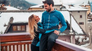 Una pareja abrazándose en un balcón con tejados nevados y montañas al fondo.