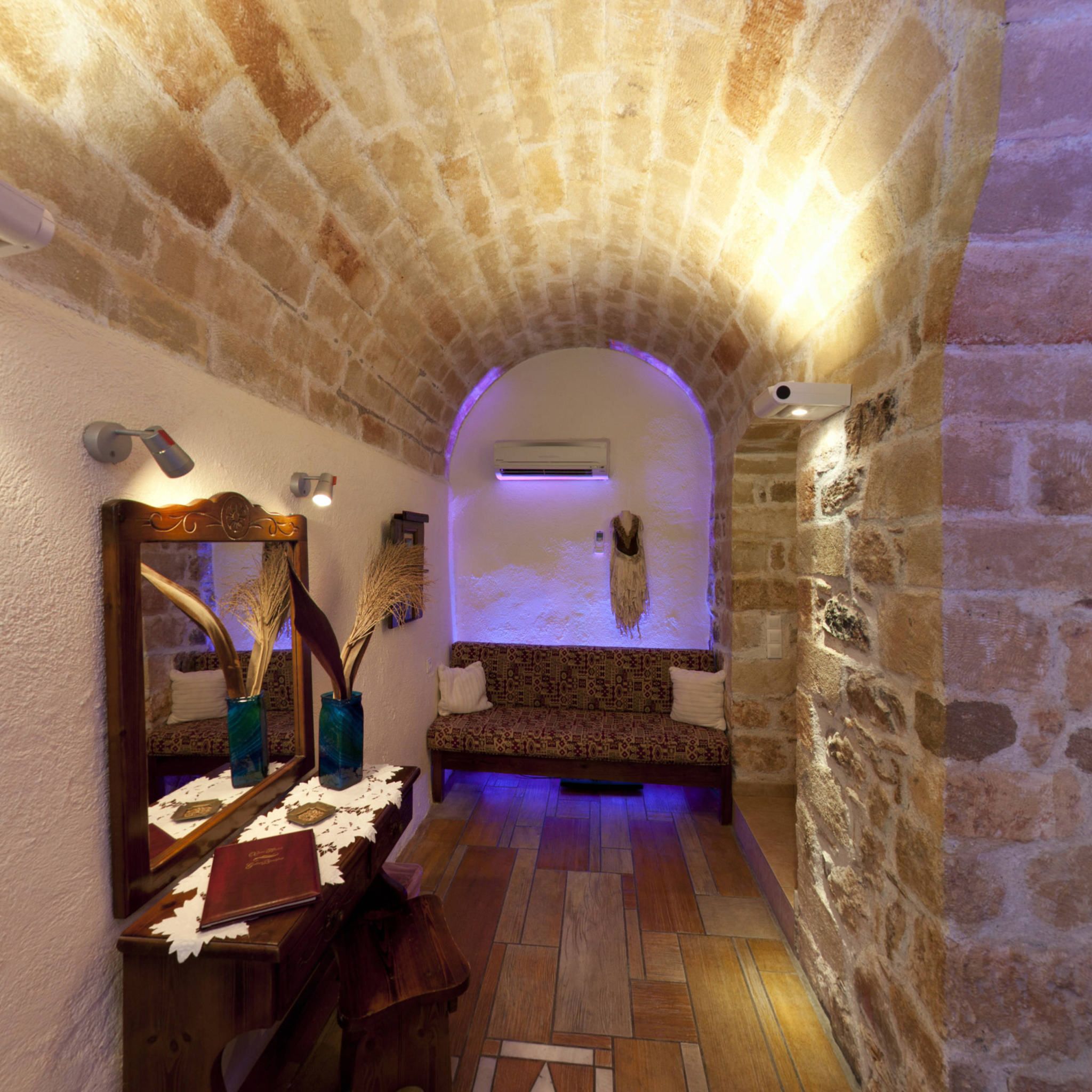 A stone-walled hallway with a wooden bench, a mirror, and a vase with dried plants.