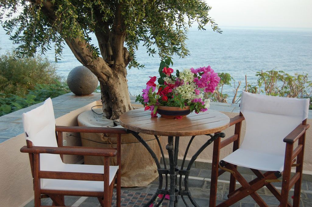 A round wooden table with a flower arrangement, two white cushioned chairs, and a large tree in the background.