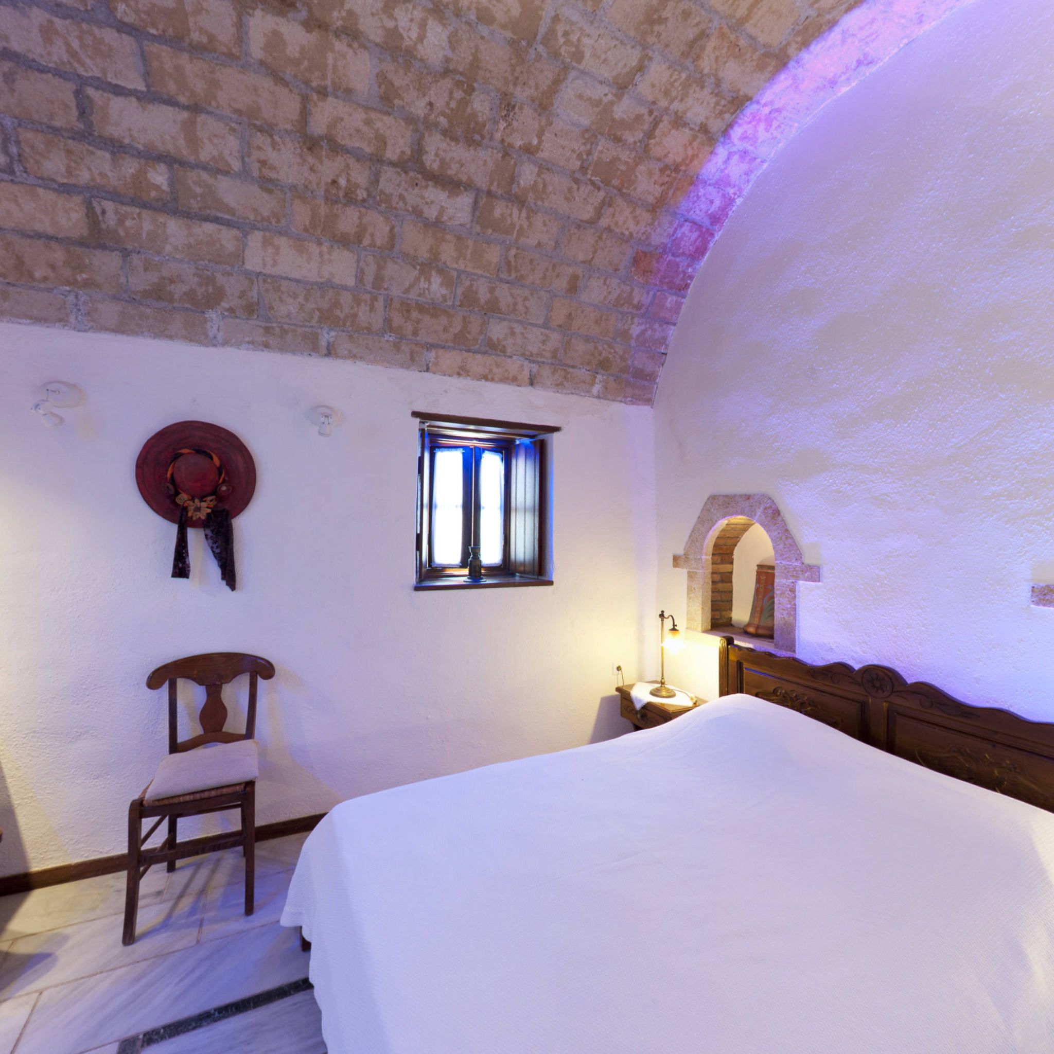 Una habitación con techo de ladrillo, una cama, una silla de madera, un ventanal pequeño y un sombrero colgado en la pared.