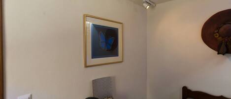 A room with a wooden desk, a framed butterfly artwork, a small shelf with a teapot, a chair, and a wall-mounted hat.
