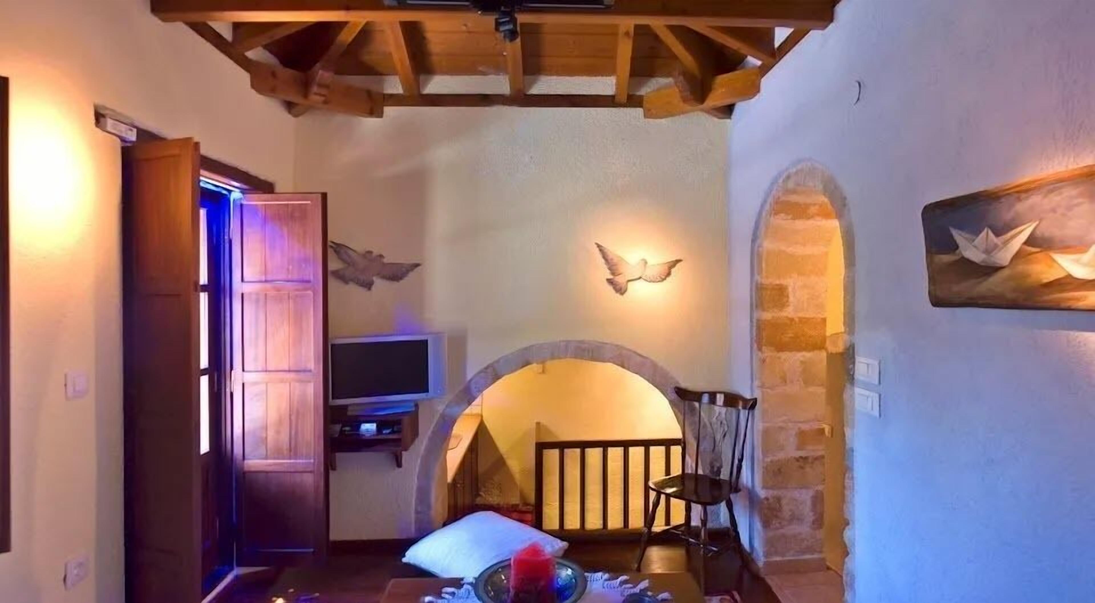 A cozy room with a wooden ceiling, a television on a stand, a bed with pillows, a small table with items, and a stone wall with a painting of birds.