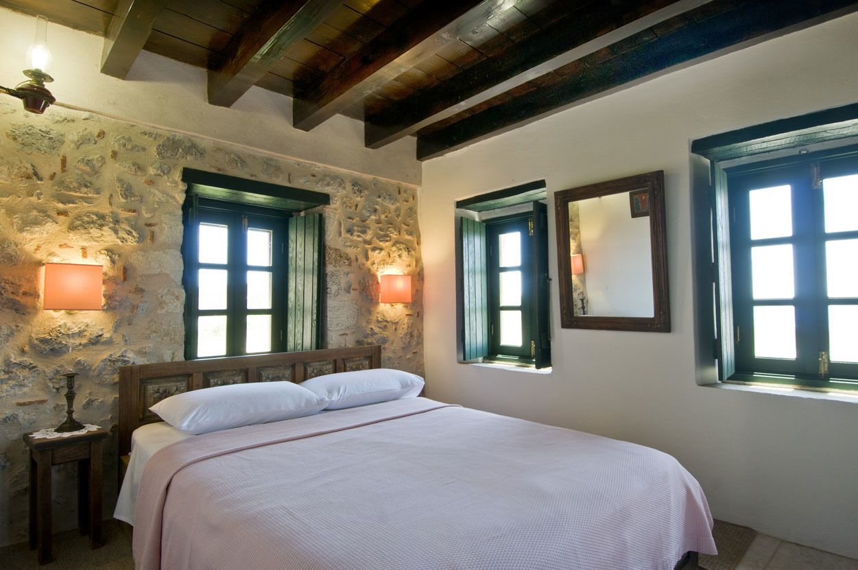 A bedroom with a stone wall, wooden beams, a bed with white linens, and two windows with shutters.