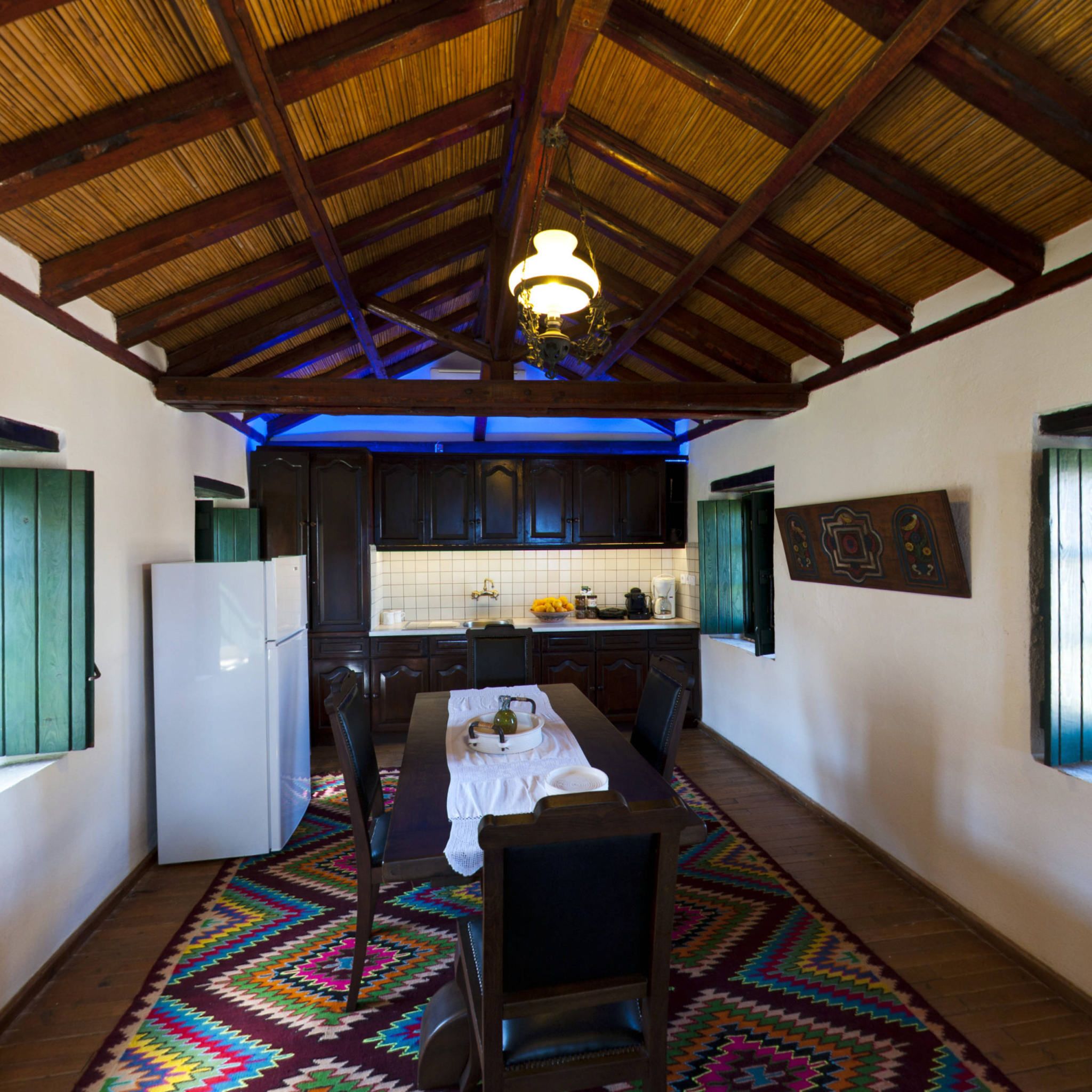 Una cocina compacta con vigas de madera, un refrigerador blanco, una mesa de comedor con sillas y una alfombra colorida.