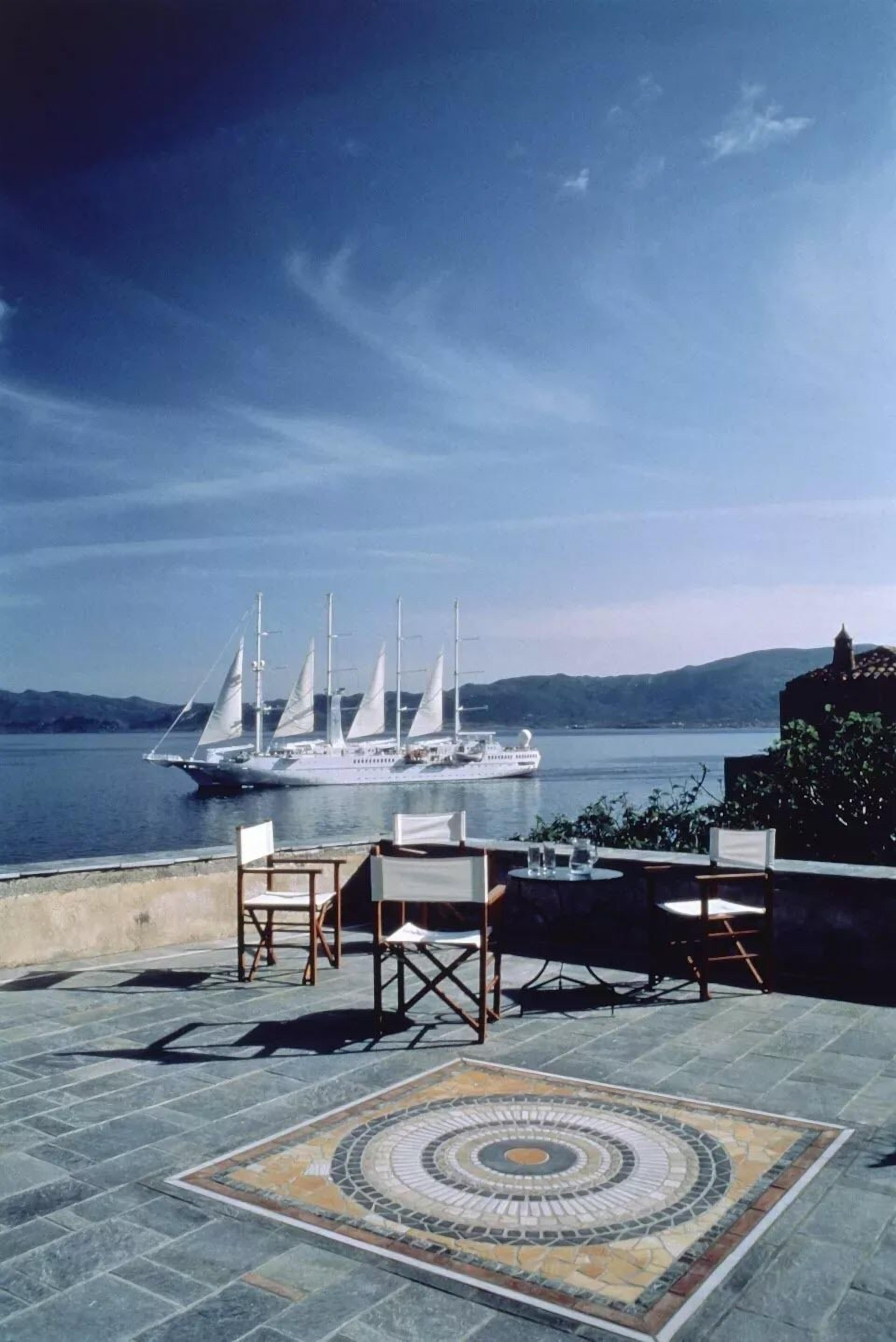 Eine Terrasse mit einem gemusterten Boden, Sitzgelegenheiten im Freien und Blick auf ein Segelboot auf dem Wasser.