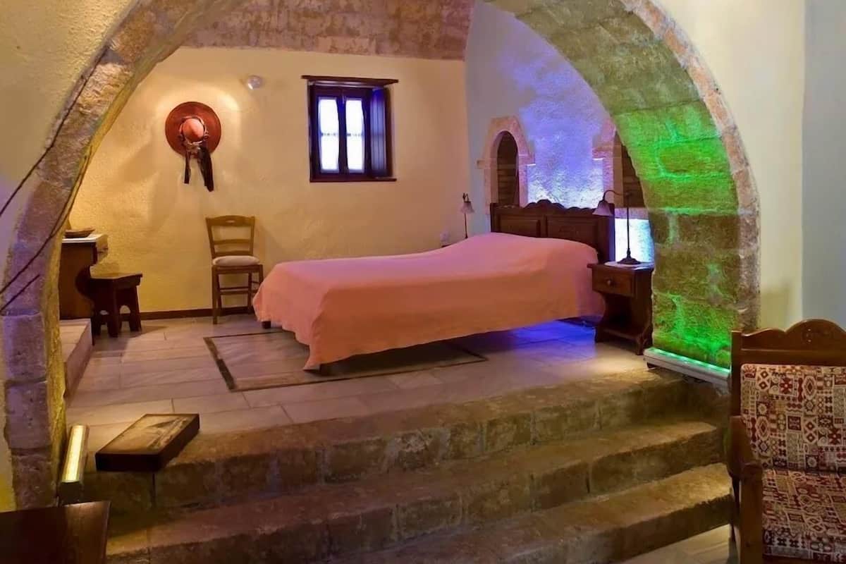 A bedroom with a stone archway, a bed, a chair, a nightstand, and a window.