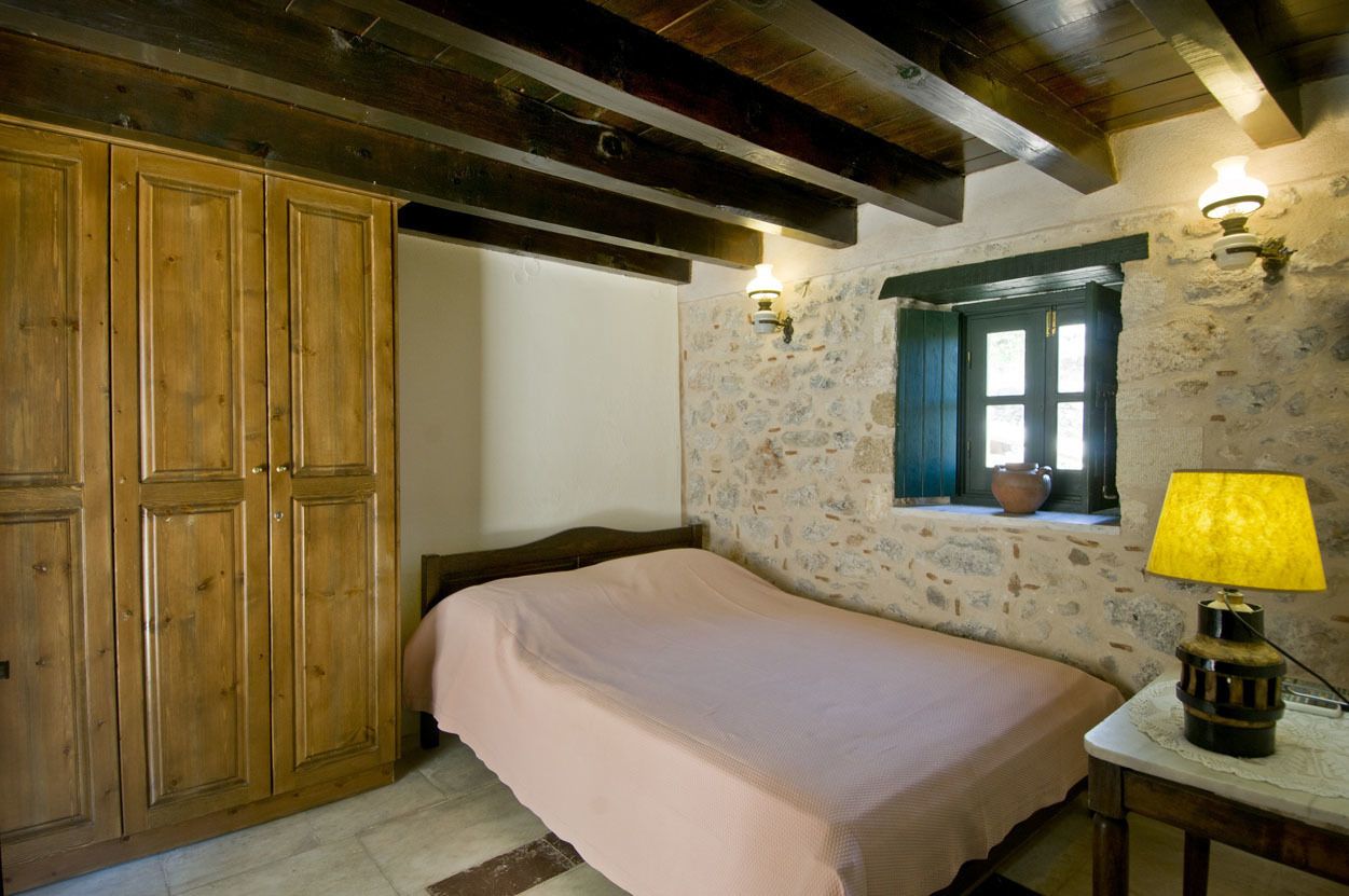 Quarto com guarda-roupa de madeira, cama com colcha rosa, parede de pedra, janela com moldura verde e mesa com abajur.