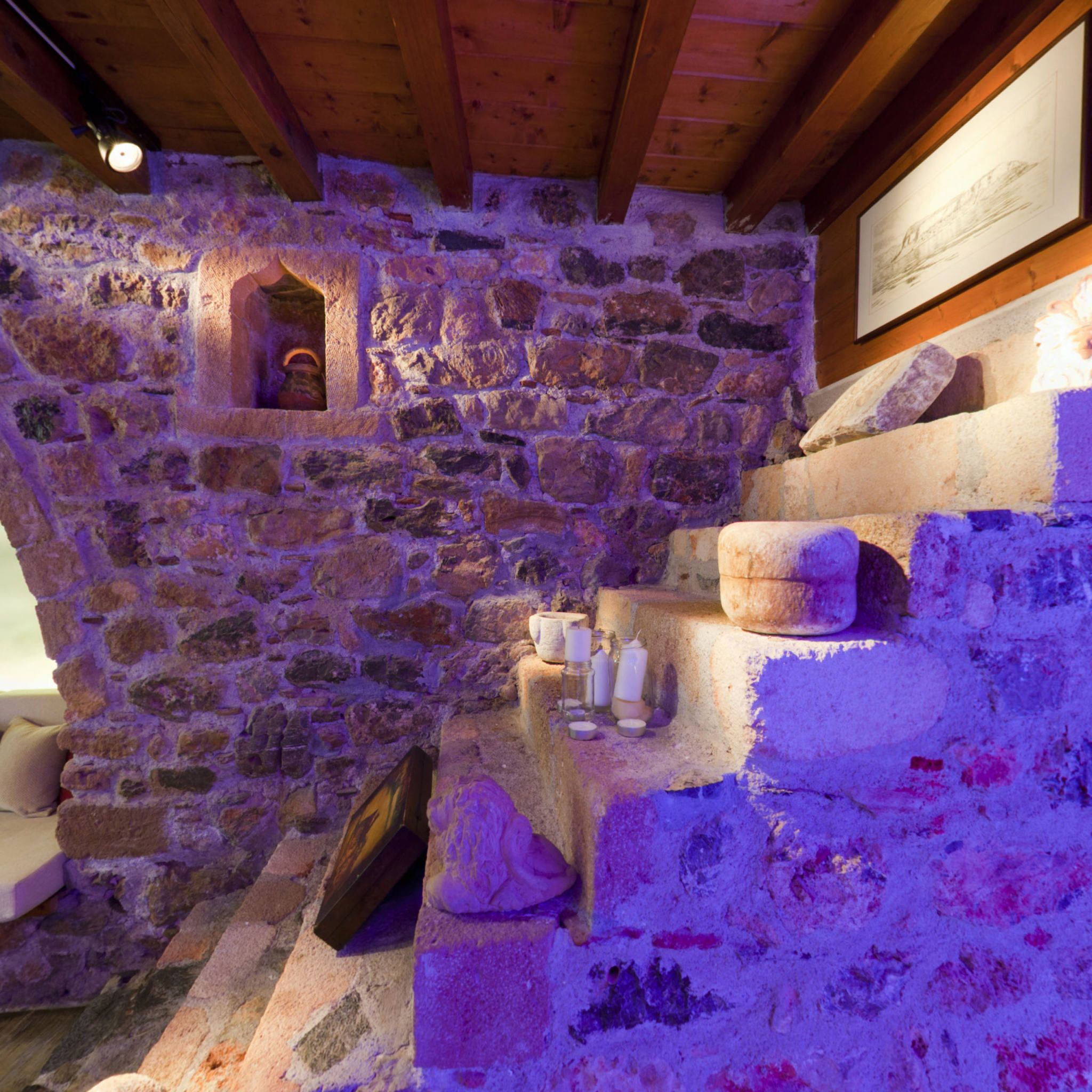 A stone-walled room with a wooden ceiling, a small alcove with a light, and a stone bench with jars.
