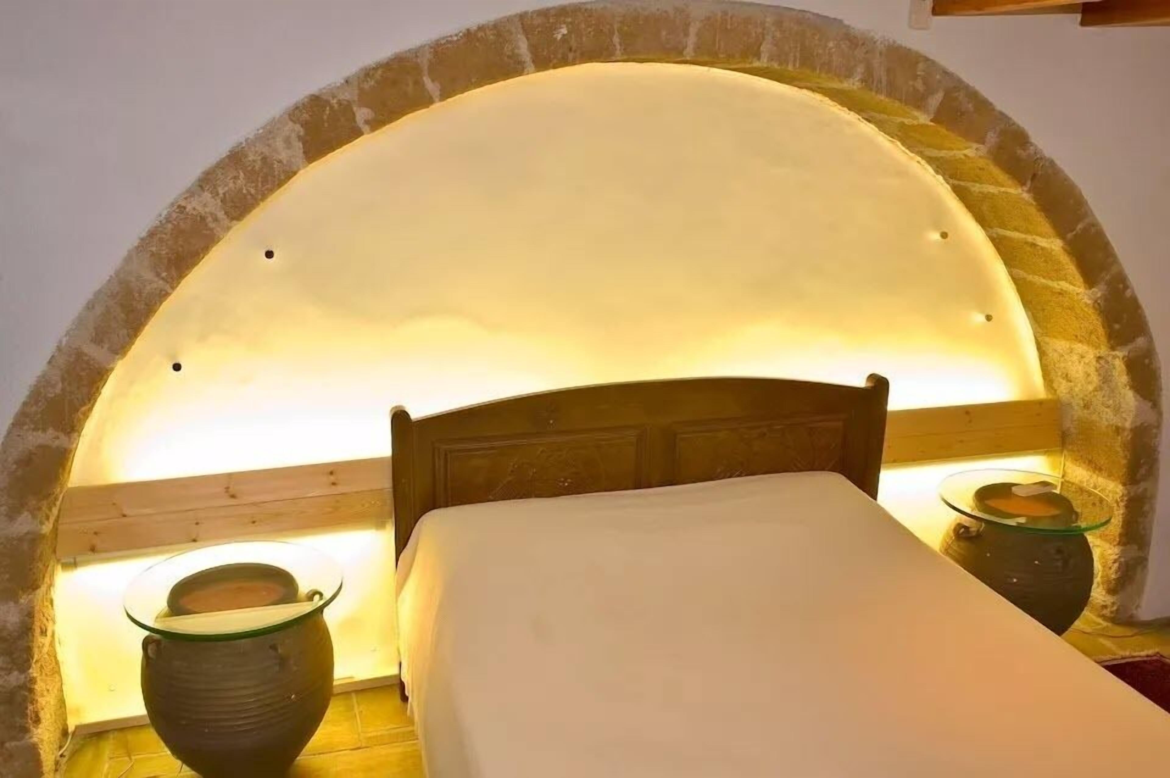 A bed with a wooden headboard under a stone archway, two bedside tables with glass bowls, and a warm light source.