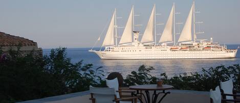 A large white cruise ship with multiple sails docked near a waterfront. There are outdoor tables and chairs with a view of the ship.