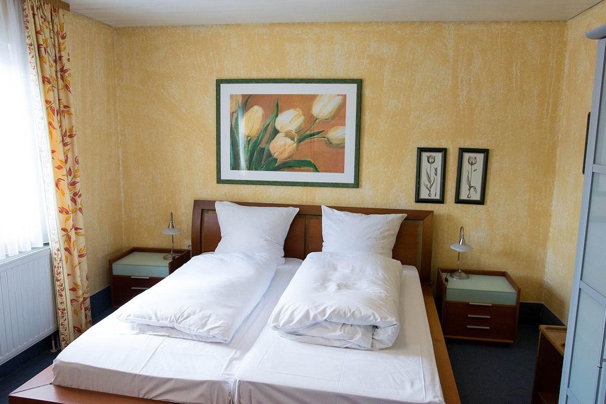 A double bed with white linens, two bedside tables, a night lamp, and a painting of tulips on the wall.